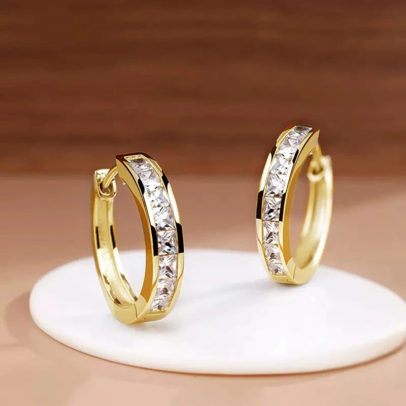 Gold hoop earrings with clear stones on a beige stand against a brown background