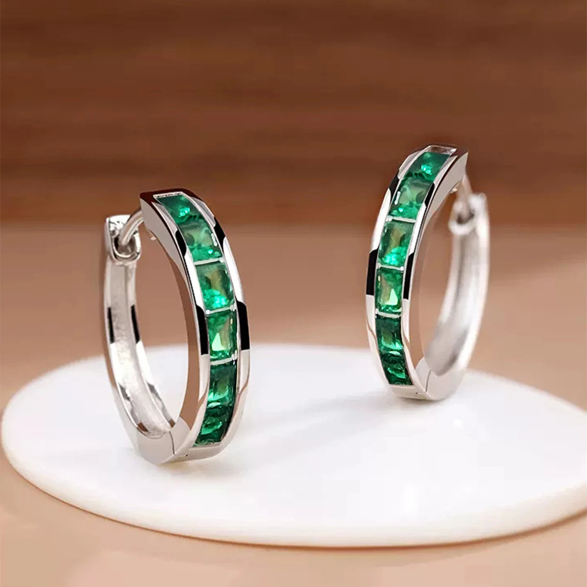 Silver hoop earrings with green gemstones on a beige stand.